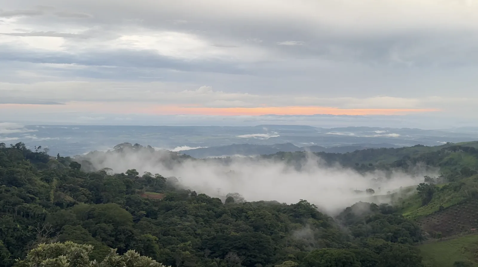 Mist drifting through green mountain valleys beneath a distant sunset viewed from Ecovilla San Mateo.